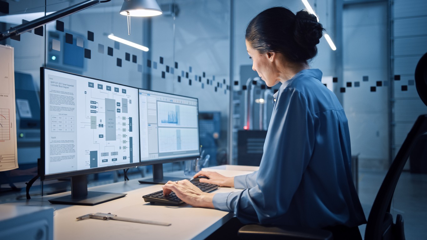 Factory Office: Portrait of Beautiful and Confident Female Industrial Engineer Working on Computer, on Screen Industrial Electronics Design Software. High Tech Facility with CNC Machinery